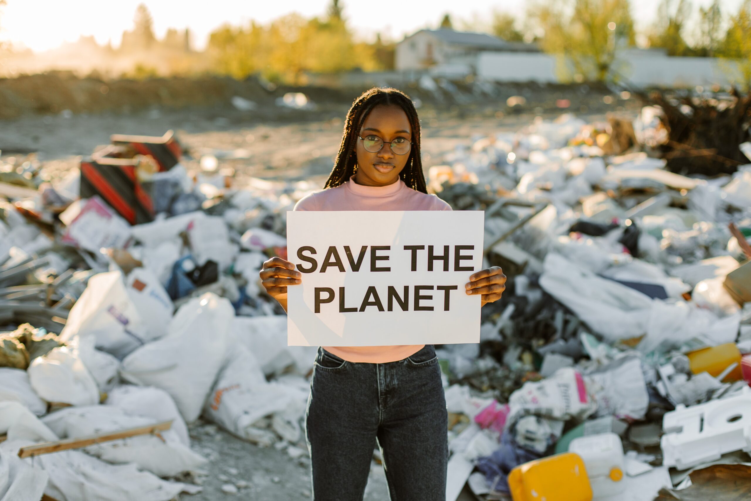 Eine Frau steht vor eine Landschaft aus Müll und hält ein Schild hoch: Save the Planet