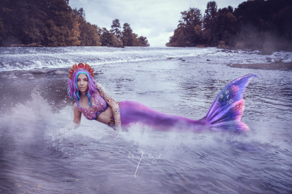 Nadine liegt im flachen Wasser auf der Seite. Sie trägt eine lila Flosse, lila Haare, einen lila BH und eine Krone.