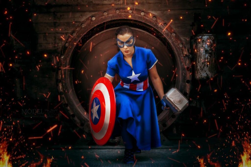Nadine trägt ein Kleid im Captain America Still. In einer Hand hält sie Thors Hammer und in der anderen Captain Americas Schild. Funken fliegen um sie her. Sie läuft geduckt.