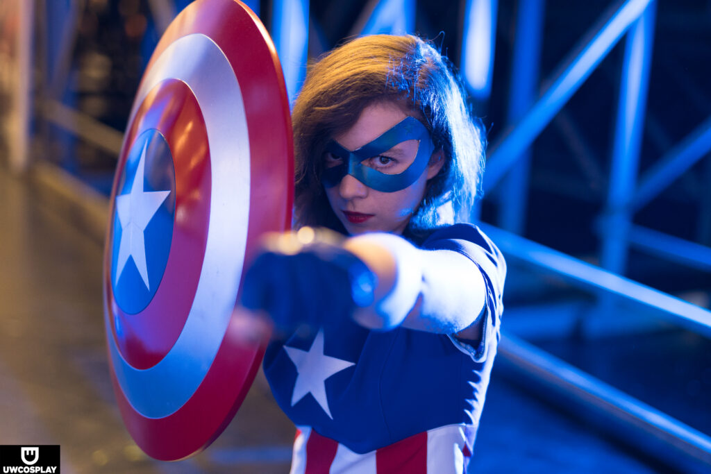 Nadine ist bis unter der Burst zu sehen. Sie tärgt ein Kleid im Captain America Stil. Sie trägt eine blaue Maske und blaue Handschuhe. EIn Faust streckt sie nach vorne. In der anderen Hand hält sie ein Schild.