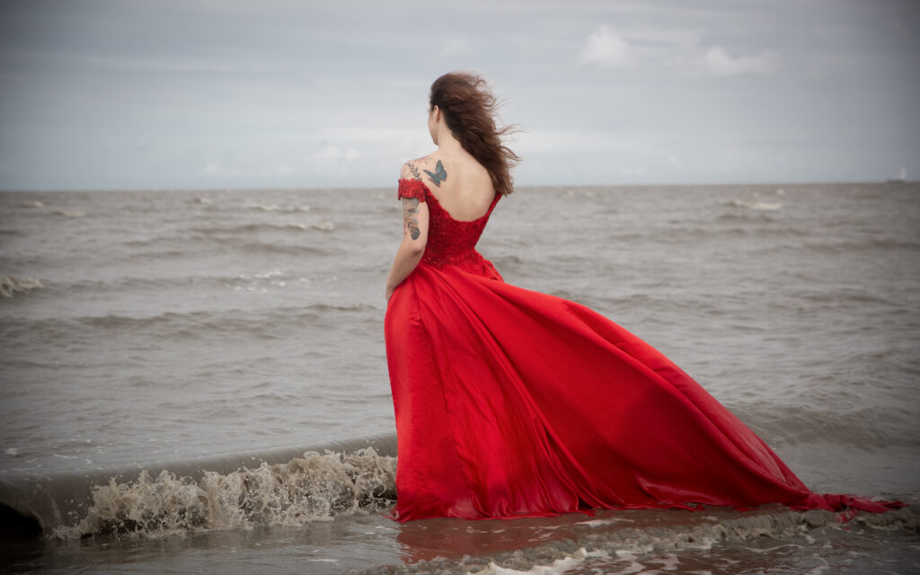 Nadine steht mit einem roten Ballkleid mit dem Rücken zur Kamera. Sie steht im Wasser am Meer. Wellen sind zu sehen.