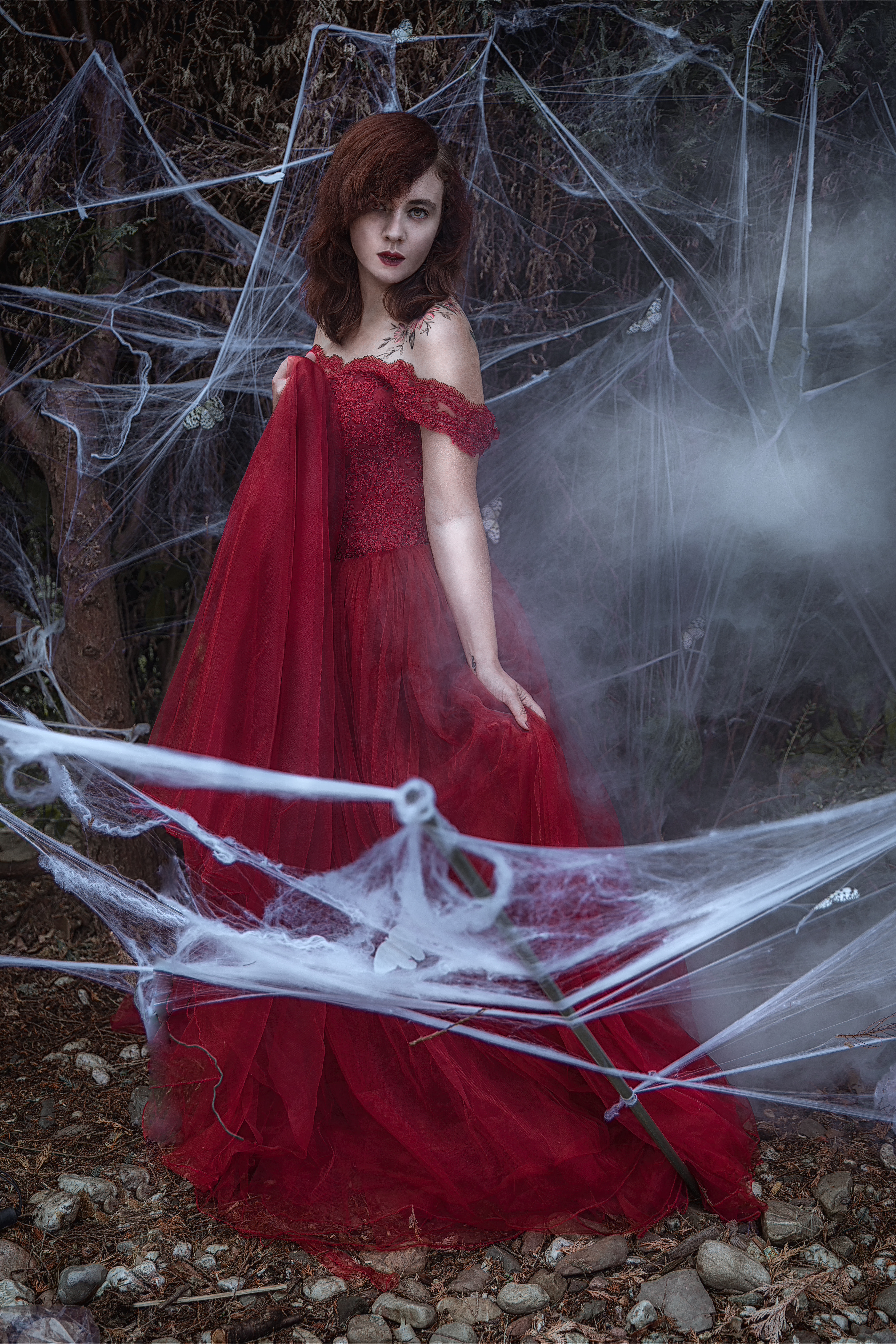 Nadine steht umgeben von Spinnennetzen in einem roten Ballkleid im freien. Ein Teil des Kleides ist in einer Hand zur Brust gezogen. Den anderen Teil des kleides hält sie in der anderen Hand leicht hoch.