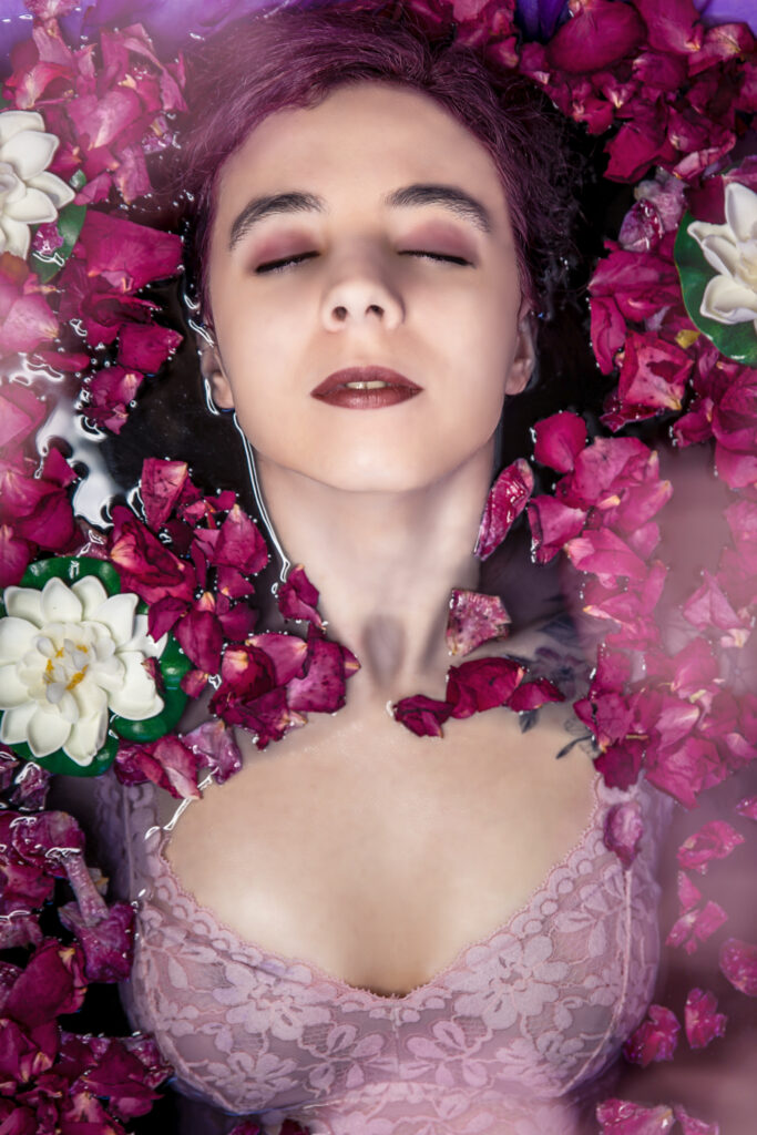 Nadine liegt mit geschlossenen Augen in einer Badewanne mit lila Wasser. Um sie herum weiße Seerosen und Rosenblüten. Sie trägt ein rosa Negligé, Man sieht sie bis zur Taille.