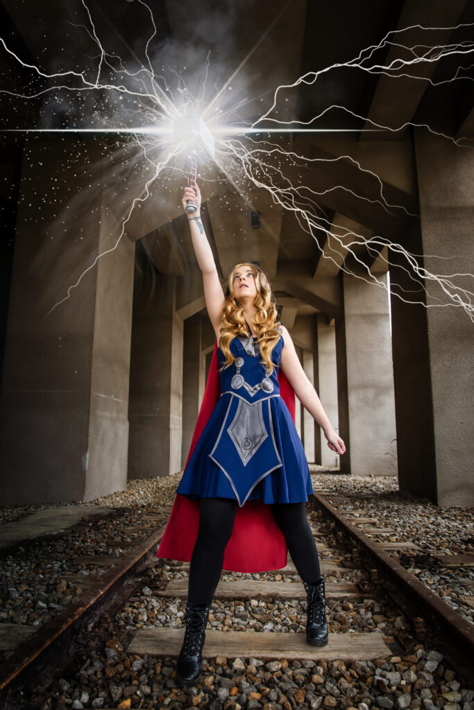 Nadine trägt ein Kleid im Thor stil. Dazu einen roten Umhang. Sie steht breitbeinig auf Schienen. Sie reckt Thors Hammer in die Luft und Blitze erscheinen. Sie hat lange blonde Haare.
