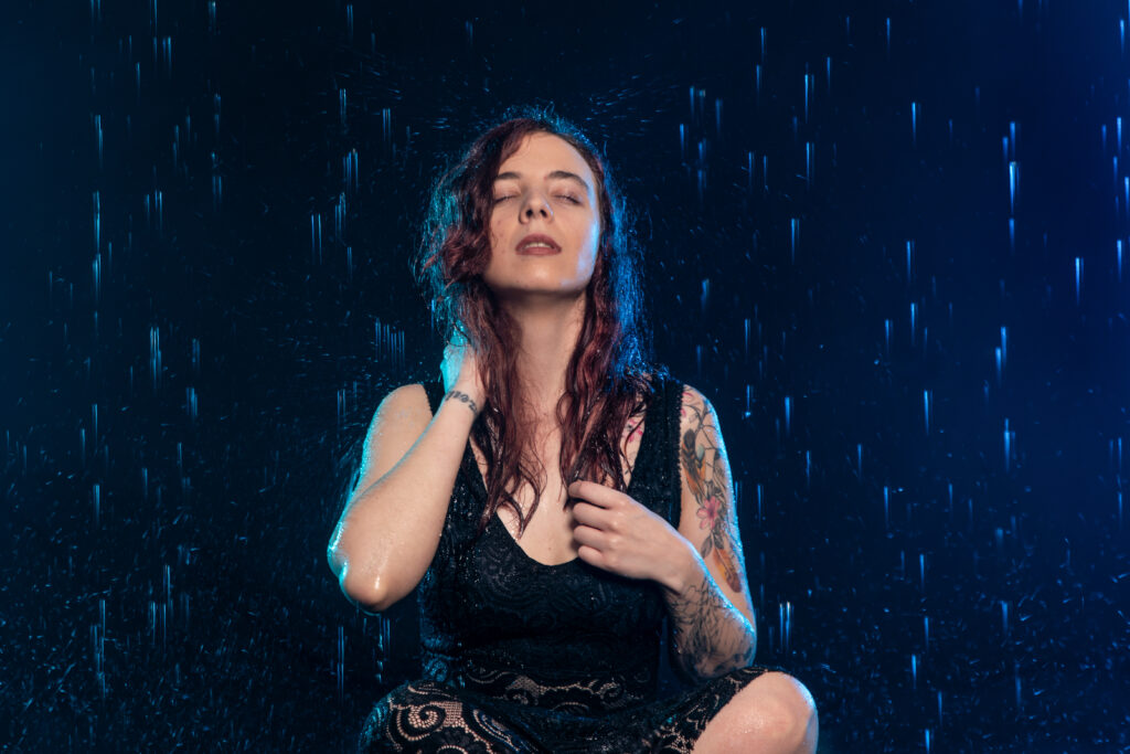Nadine trägt ein schwarzes Kleid und sitzt in einem Studio mit Regen. Eine Hand an ihren Haarspitzen und eine Hand etwas höher. Der Kopf ist in den Nacken gelegt und die Augen geschlossen während die Tropfen fallen.