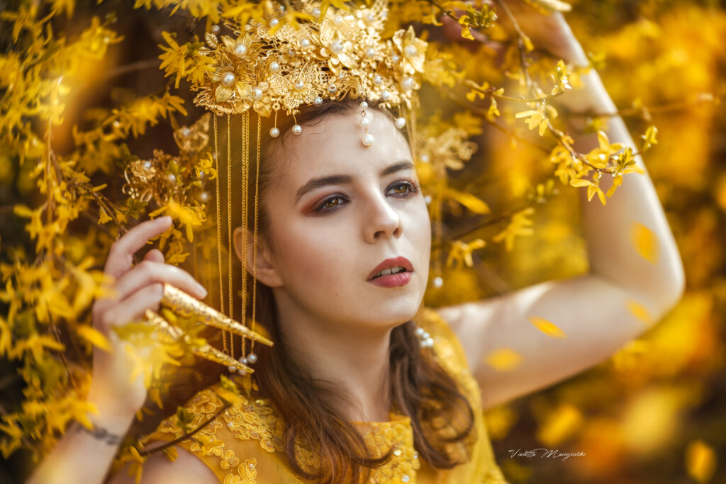 Nadine trägt ein goldenes Kleid, einen goldenen Kopfschmuck und goldene Krallen über den Fingerspitzen. Sie lehnt an einen Buch mit gelben Blüten. Sie ist bis zur Brust zu sehen. 