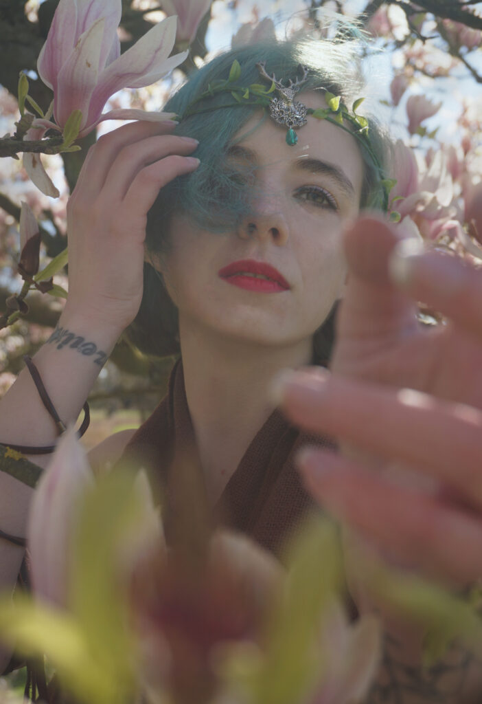Nadine steht in einem Kirschblüten Baum. Sie ist bis zur Brust zu sehen. Zweige und Blüten umgeben sie. Sie hat grüne Haare und einen Kopfschmuck auf. Eine Hand ist Richtung Kamera gestreckt. Die andere an ihrem Schlüsselbein. 
