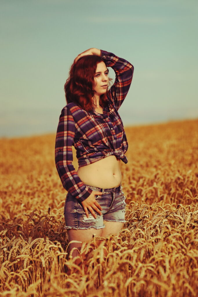 Nadine steht in einem Weizenfeld.Sie trägt eine kurze Jeans und hat eine blau-rot karrierte Bluse bauchfrei zusammengebunden.