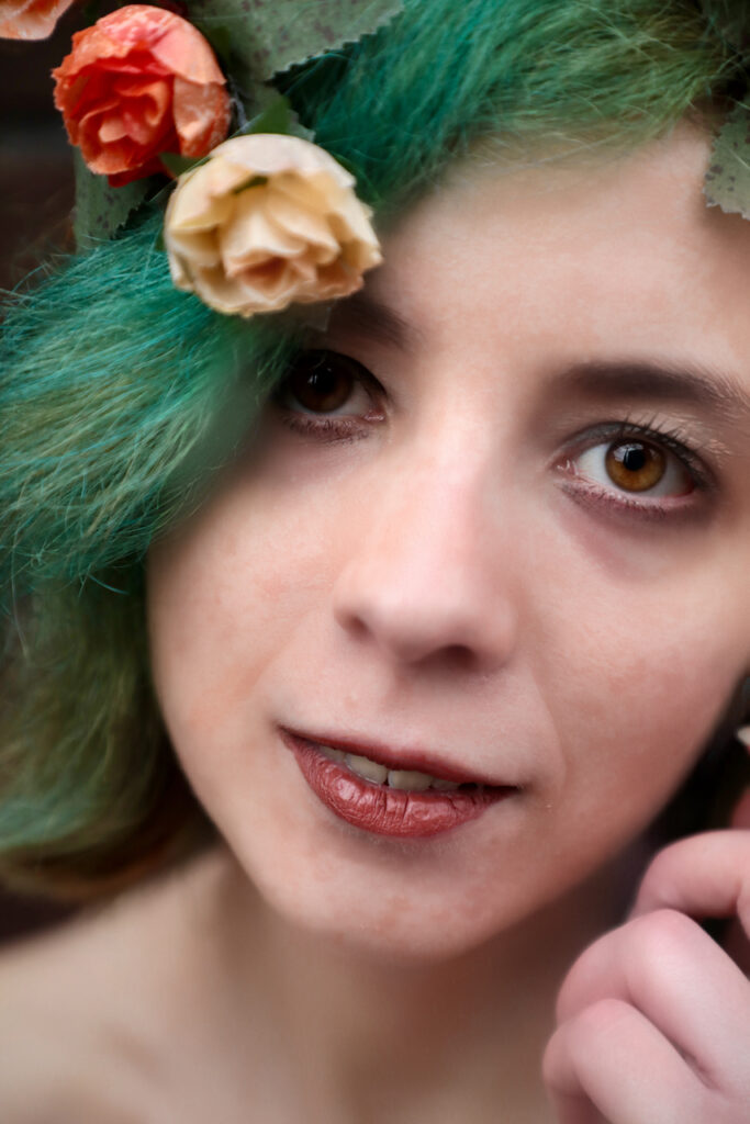 Eine Gesichtsaufnahme von Nadine. Sie hat große braune Augen und grüne kurze Haare. man sieht orangene Blumen im Haar. Eine Hand ist in der Nähe der Wange