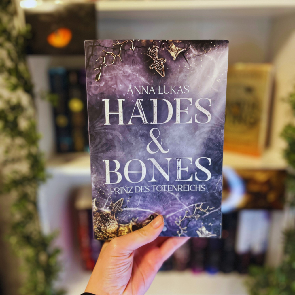 Ein Buch wird in der Hand gehalten. Das Cover ist zu sehen. Der Titel des Buches lautet "HADES & BONES - Prinz des Totenreichs" und der Autorenname "Anna Lukas" ist oben auf dem Cover zu sehen. Das Cover hat einen dunklen, nebligen Hintergrund mit lila und blauen Tönen. Es gibt auch einige silberne Schmuckelemente wie eine Kette und Anhänger, die wie ein Kreuz und ein Knochen aussehen. Im Hintergrund ist ein unscharfes Bücherregal mit verschiedenen Büchern zu erkennen.