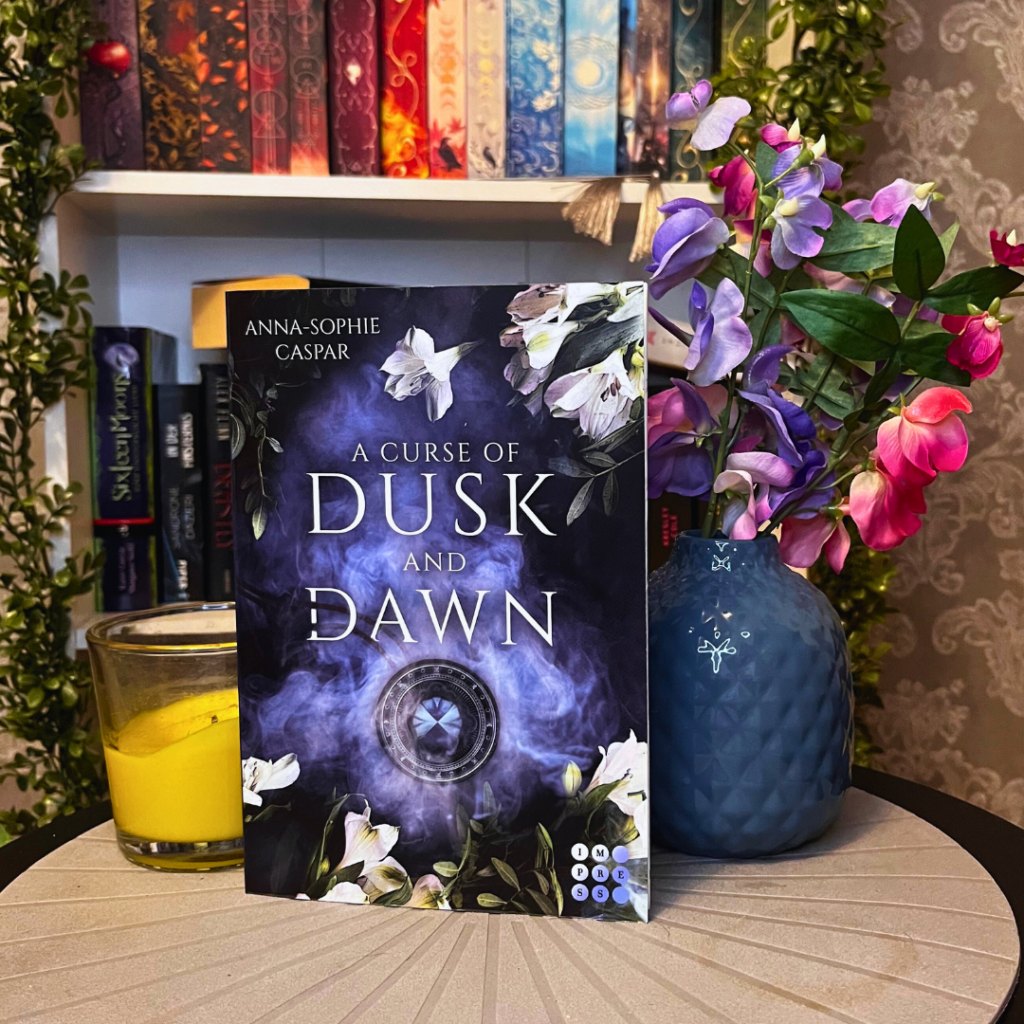 Das Bild zeigt ein Buch mit dem Titel "A Curse of Dusk and Dawn" von Anna-Sophie Caspar. Das Cover des Buches ist in dunklen Farbtönen gehalten, mit einem Design aus weißen Blumen und lila Rauchschwaden. Im Hintergrund ist ein Bücherregal mit vielen bunten Bücherrücken zu sehen. Auf der linken Seite des Bildes befindet sich eine gelbe Kerze in einem Glas. Rechts neben dem Buch steht eine blaue Vase mit einer Pflanze, die lila und rosa Blüten trägt. Das Buch und die Gegenstände stehen auf einer grauen Oberfläche mit kreisförmigen Mustern.