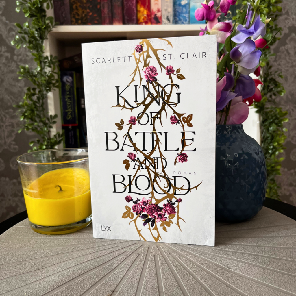 Ein Buch mit dem Titel "King of Battle and Blood" von Scarlett St. Clair, das auf einem Tisch steht. Der Bucheinband ist hauptsächlich weiß mit einer stilisierten Darstellung von Ranken und Blumen in Gold und Rosa. Links neben dem Buch befindet sich eine gelbe Kerze in einem Glasbehälter. Rechts steht eine dunkelblaue Vase mit einer Mischung aus künstlichen lila und rosa Blumen. Im Hintergrund sind unscharf weitere Bücher und eine grüne Pflanze zu erkennen. Der Tisch hat eine strukturierte Oberfläche mit radialen Linien.