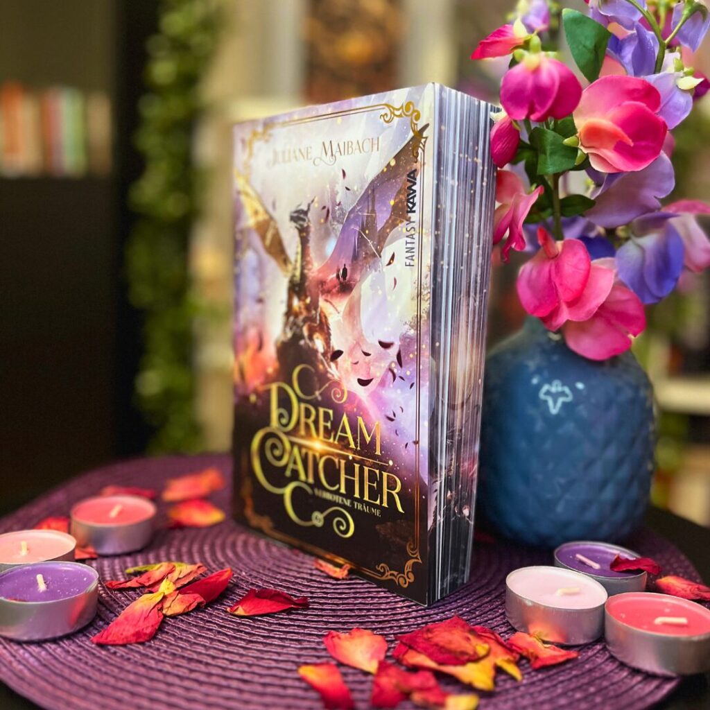 Das Bild zeigt ein aufrecht stehendes Buch mit dem Titel "Dream Catcher" von Juliane Maibach. Das Cover des Buches ist farbenfroh mit einer magischen Szenerie, die eine Fee zeigt, die in der Nähe eines großen, leuchtenden Traumfängers schwebt. Neben dem Buch befindet sich eine Vase mit lebhaften, bunten Blumen und einige brennende Teelichter in verschiedenen Farben sind auf einem dunkelvioletten Tisch verteilt. Um die Kerzen herum liegen verstreut rote und orange Blütenblätter. Der Hintergrund ist unscharf, aber man kann erkennen, dass sich weitere Bücher und Pflanzen im Raum befinden.