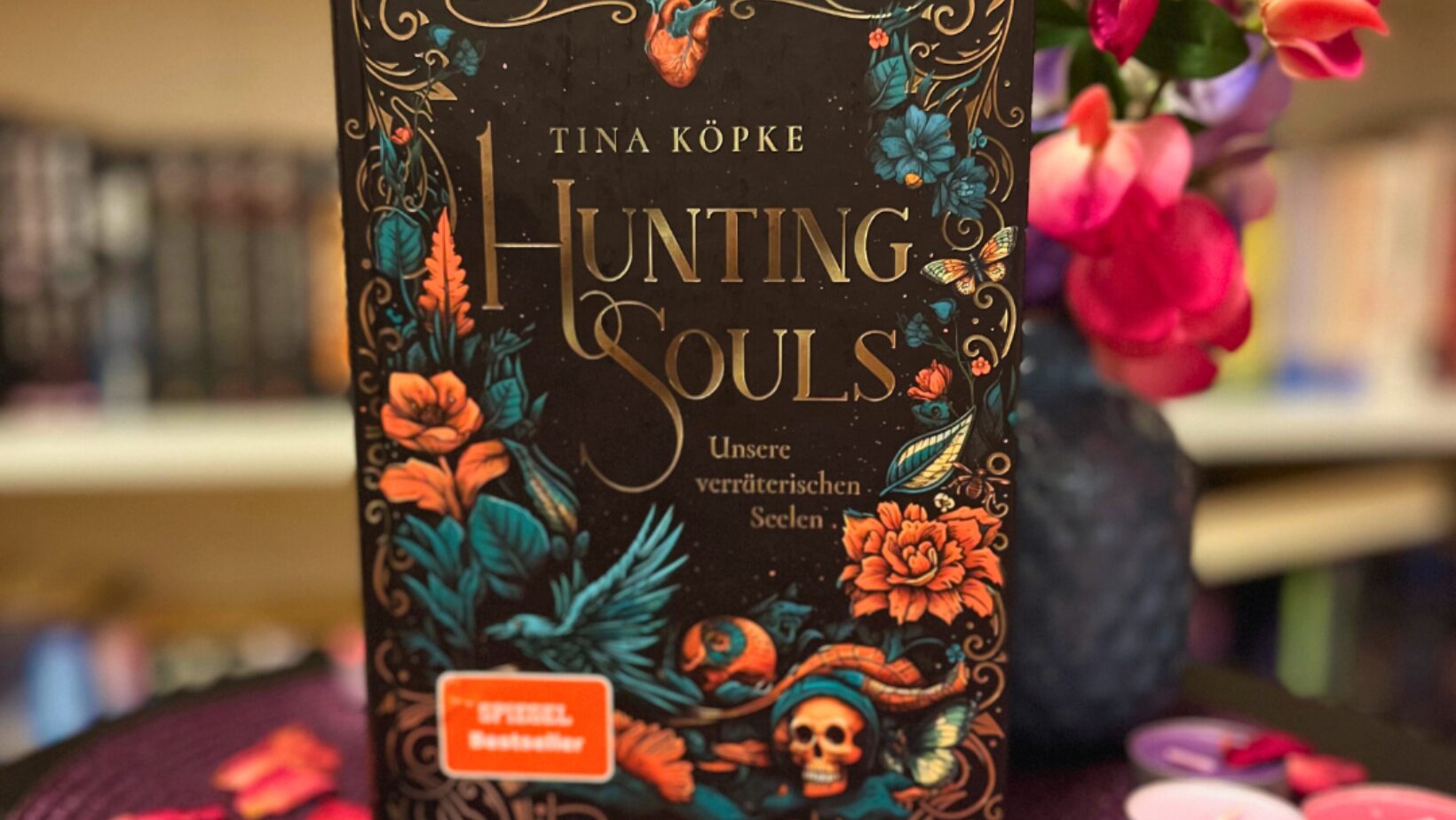 Das Bild zeigt ein Buch mit dem Titel "Hunting Souls - Unsere verräterischen Seelen" von Tina Köpke. Das Buchcover ist reich verziert mit einem dunklen Hintergrund und einem floralen Muster in leuchtenden Farben wie Orange, Blau und Rot. Einige Elemente wie ein Herz und ein Schädel sind ebenfalls abgebildet. Das Buch steht auf einem runden, violetten Tischset, umgeben von einigen brennenden Kerzen in Rosa und Rot sowie verstreuten Blütenblättern. Im Hintergrund ist unscharf ein Bücherregal zu erkennen.