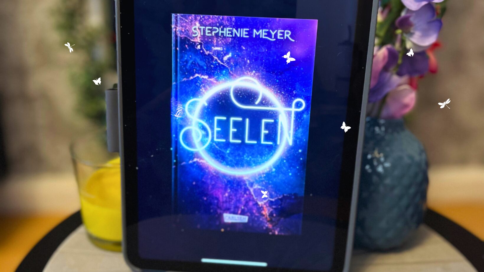 Auf dem Bild ist ein Tablet zu sehen, das auf einem runden, hellen Holztisch steht. Auf dem Bildschirm des Tablets ist das Cover eines Buches mit dem Titel "Seelen" von Stephenie Meyer sichtbar. Das Cover zeigt einen leuchtenden, blauen und lila Nebel im Hintergrund mit einem leuchtenden, weißen Kreis, der wie eine stilisierte Darstellung einer Seele aussieht. Neben dem Tablet befindet sich eine Vase mit lila und rosa Blumen und ein Glas mit einer gelben Flüssigkeit, vermutlich Saft. Der Hintergrund ist unscharf, aber es scheint eine Pflanze und eine Wand zu geben.