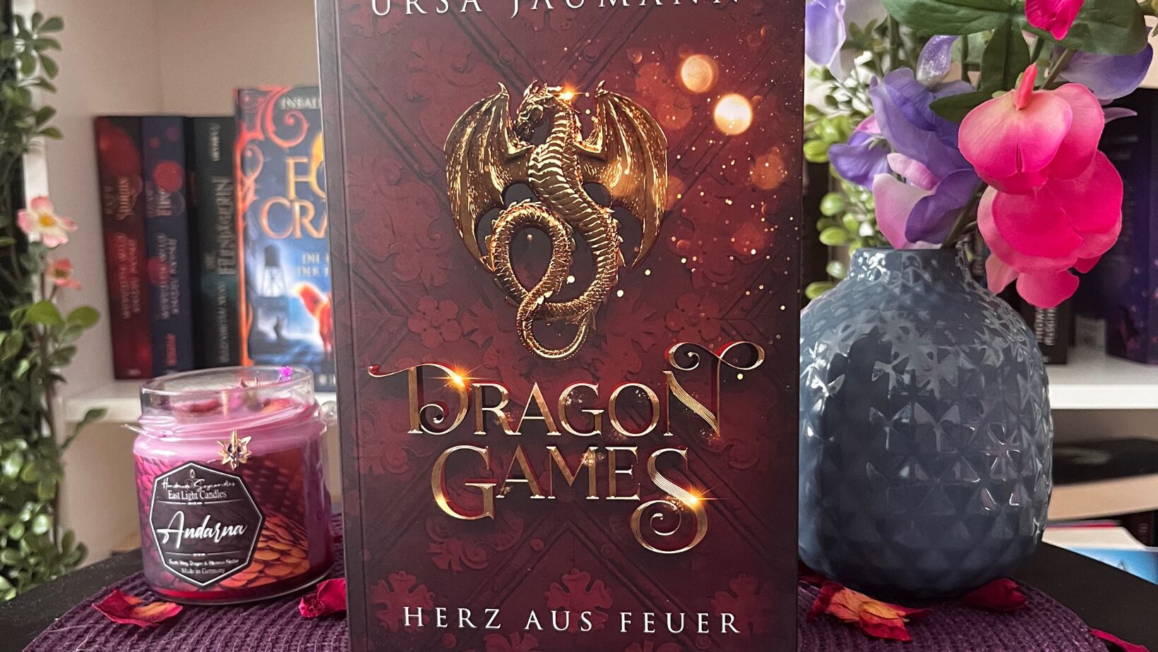 Das Bild zeigt ein Buch mit dem Titel "Dragon Games: Herz aus Feuer" von Ursa Jaumann. Das Cover des Buches ist in Rottönen gehalten und zeigt einen goldenen Drachen, der sich um ein Schwert windet. Das Buch steht auf einem runden, lila geflochtenen Untersetzer, der mit getrockneten Rosenblättern und kleinen lila Teelichtern dekoriert ist. Im Hintergrund sind unscharf weitere Bücher und eine Vase mit bunten Blumen zu sehen.