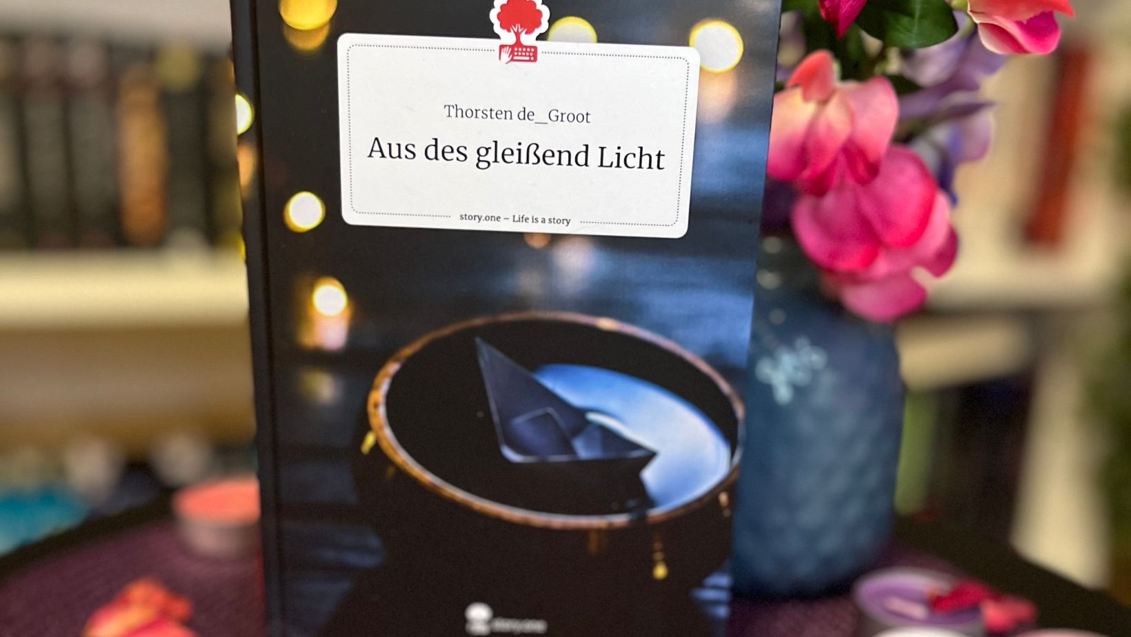 Das Bild zeigt ein Buch mit dem Titel "Aus des gleißend Licht" von Thorsten de Groot. Das Buch steht aufrecht auf einem Tisch, der mit einem lila Tuch bedeckt ist. Um das Buch herum liegen getrocknete Rosenblätter und mehrere Teelichter in verschiedenen Farben. Im Hintergrund sind unscharf Bücherregale und eine Vase mit rosa und lila Blumen zu sehen.