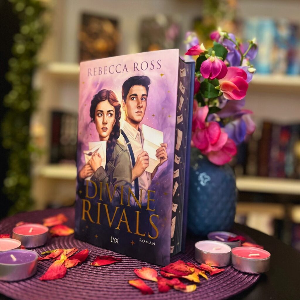 Das Bild zeigt ein Buch mit dem Titel "Divine Rivals" von Rebecca Ross. Das Cover des Buches zeigt zwei gezeichnete Figuren, eine Frau und einen Mann, die beide Briefe in den Händen halten. Der Hintergrund des Covers ist lila mit goldenen Sternen. Das Buch steht auf einem runden Tisch, der mit einem lila Tuch bedeckt ist. Um das Buch herum liegen getrocknete Rosenblätter und mehrere Teelichter in verschiedenen Farben (rosa, lila und weiß). Im Hintergrund sind unscharf Bücherregale und eine Vase mit rosa und lila Blumen zu sehen.