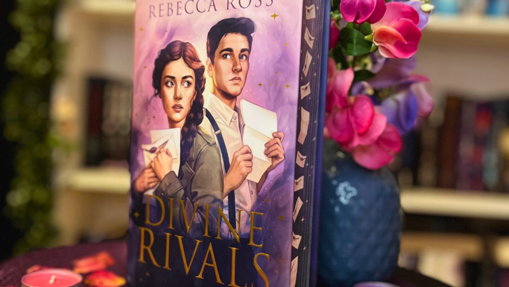 Das Bild zeigt ein Buch mit dem Titel "Divine Rivals" von Rebecca Ross. Das Cover des Buches zeigt zwei gezeichnete Figuren, eine Frau und einen Mann, die beide Briefe in den Händen halten. Der Hintergrund des Covers ist lila mit goldenen Sternen. Das Buch steht auf einem runden Tisch, der mit einem lila Tuch bedeckt ist. Um das Buch herum liegen getrocknete Rosenblätter und mehrere Teelichter in verschiedenen Farben (rosa, lila und weiß). Im Hintergrund sind unscharf Bücherregale und eine Vase mit rosa und lila Blumen zu sehen.