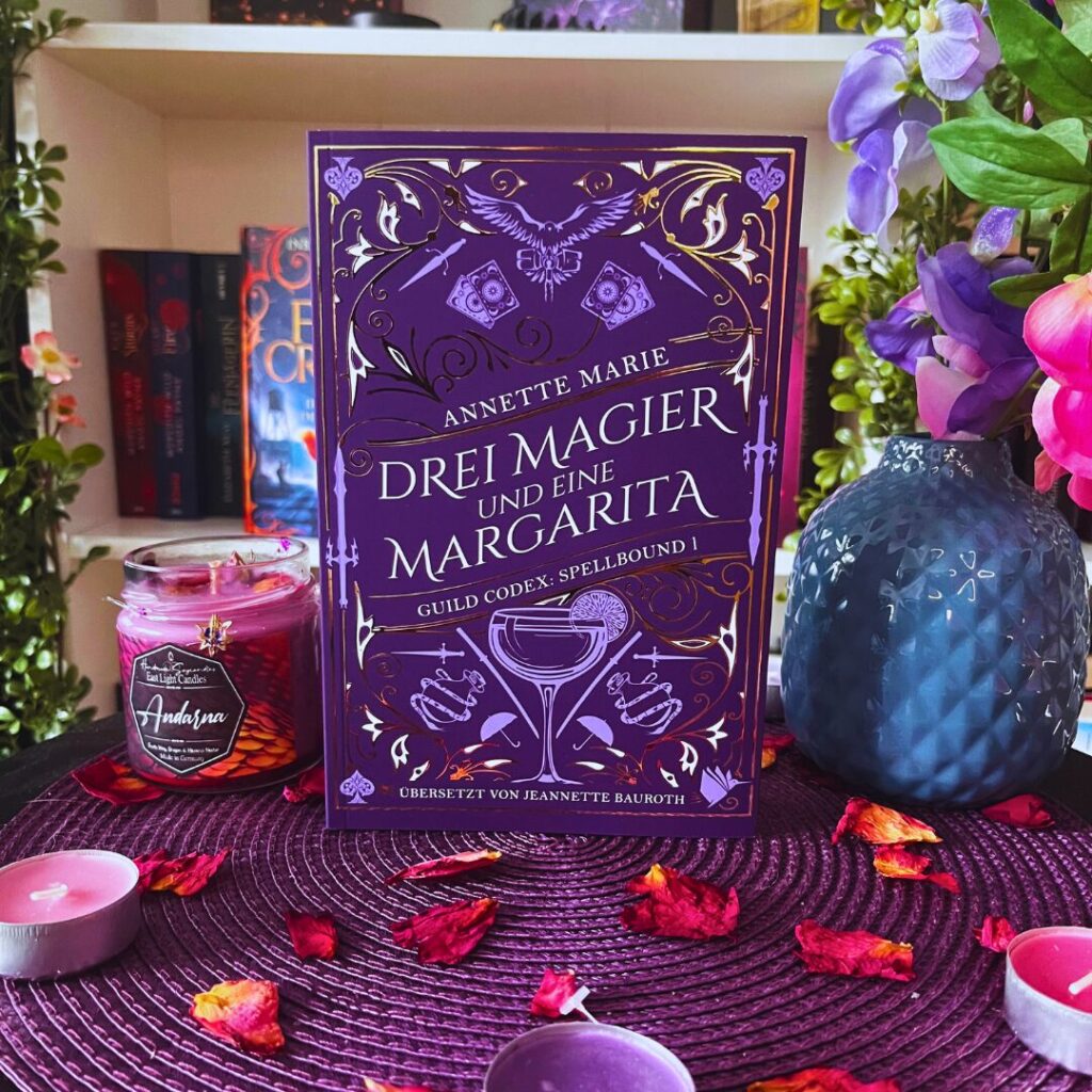 Das Bild zeigt ein Buch mit dem Titel "Drei Magier und eine Margarita" von Annette Marie. Es ist das erste Buch der Reihe "Guild Codex: Spellbound". Das Buch hat ein auffälliges lila Cover mit goldenen Verzierungen und Illustrationen, darunter ein Cocktailglas und magische Symbole. Es steht auf einem lila Untergrund, umgeben von lila Teelichtern und getrockneten Blütenblättern. Links neben dem Buch steht eine lila Kerze im Glas mit der Aufschrift "Andarna". Rechts vom Buch befindet sich eine blaue Vase mit lila und rosa Blumen. Im Hintergrund sind Bücherregale mit weiteren Büchern und Pflanzen zu sehen.