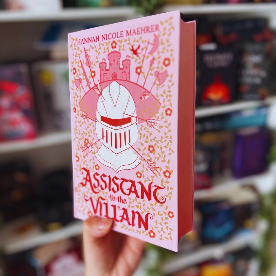 Das Bild zeigt eine Hand, die ein Buch hält. Das Buch hat einen rosa Einband und trägt den Titel "Assistant to the Villain" von Hannah Nicole Maehrer. Auf dem Cover ist eine stilisierte Ritterrüstung mit einem Visier abgebildet, umgeben von Pfeilen und einer Burg im Hintergrund. Das Design ist von floralen Mustern umgeben. Im Hintergrund sieht man ein Bücherregal mit verschiedenen anderen Büchern, die in unterschiedlichen Farben und Designs angeordnet sind.