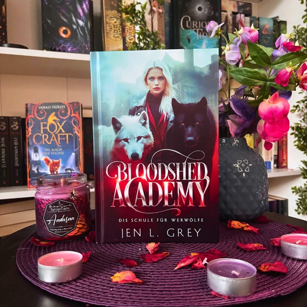 Das Bild zeigt ein Buch mit dem Titel "Bloodshed Academy: Die Schule für Werwölfe" von Jen L. Grey. Auf dem Buchcover ist eine blonde Frau abgebildet, die von einem weißen und einem schwarzen Wolf flankiert wird. Das Buch steht auf einem runden, lila Untersetzer, umgeben von getrockneten Blütenblättern und mehreren kleinen Kerzen. Links neben dem Buch steht eine violette Kerze im Glas mit der Aufschrift "Andarna". Im Hintergrund sind Bücherregale mit verschiedenen Büchern zu sehen, darunter ein Buch mit dem Titel "Fox Craft: Die Magie der Füchse". Rechts neben dem Buch steht eine Vase mit bunten Blumen.