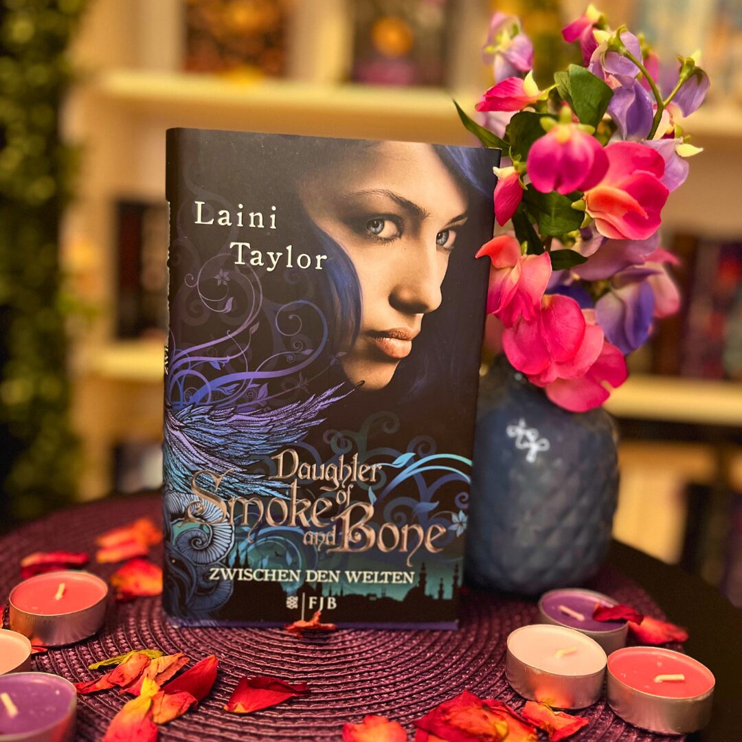 Das Bild zeigt das Buch "Daughter of Smoke and Bone" von Laini Taylor. Das Buch steht auf einem runden, lila Untersetzer, der mit roten Rosenblättern und kleinen Teelichtern in verschiedenen Farben dekoriert ist. Rechts neben dem Buch steht eine Vase mit bunten Blumen in Pink und Lila. Im Hintergrund sind unscharf Bücherregale zu erkennen.