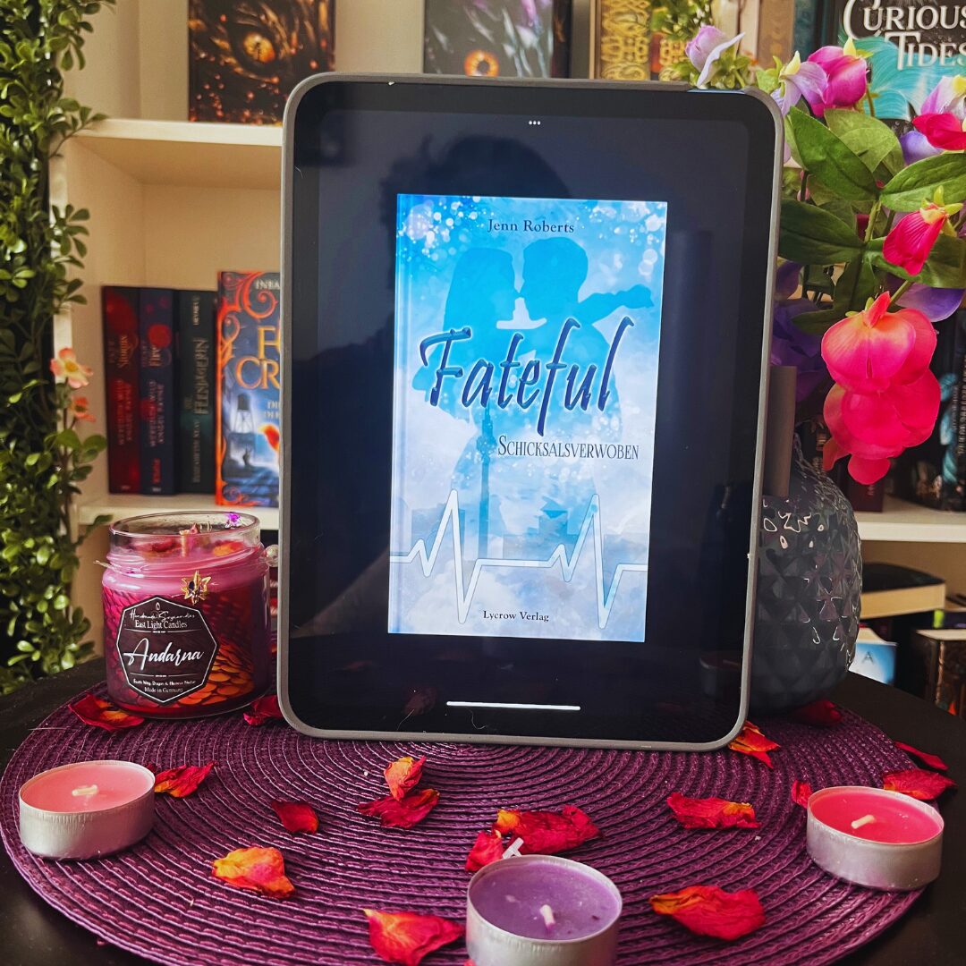 Das Bild zeigt ein Tablet, das auf einem runden Tisch steht. Auf dem Bildschirm des Tablets ist das Cover eines Buches mit dem Titel "Fateful: Schicksalsverwoben" von Jenn Roberts zu sehen. Das Cover ist in Blautönen gehalten und zeigt zwei Silhouetten, die sich gegenüberstehen, sowie eine Herzschlaglinie am unteren Rand. Der Tisch ist mit einem lila Platzdeckchen bedeckt, auf dem mehrere kleine Teelichter und getrocknete Rosenblätter verteilt sind. Links neben dem Tablet steht eine rote Kerze im Glas mit dem Etikett "Andarna". Rechts neben dem Tablet steht eine Vase mit pinken und lila Blumen. Im Hintergrund sind Bücherregale mit verschiedenen Büchern zu sehen.