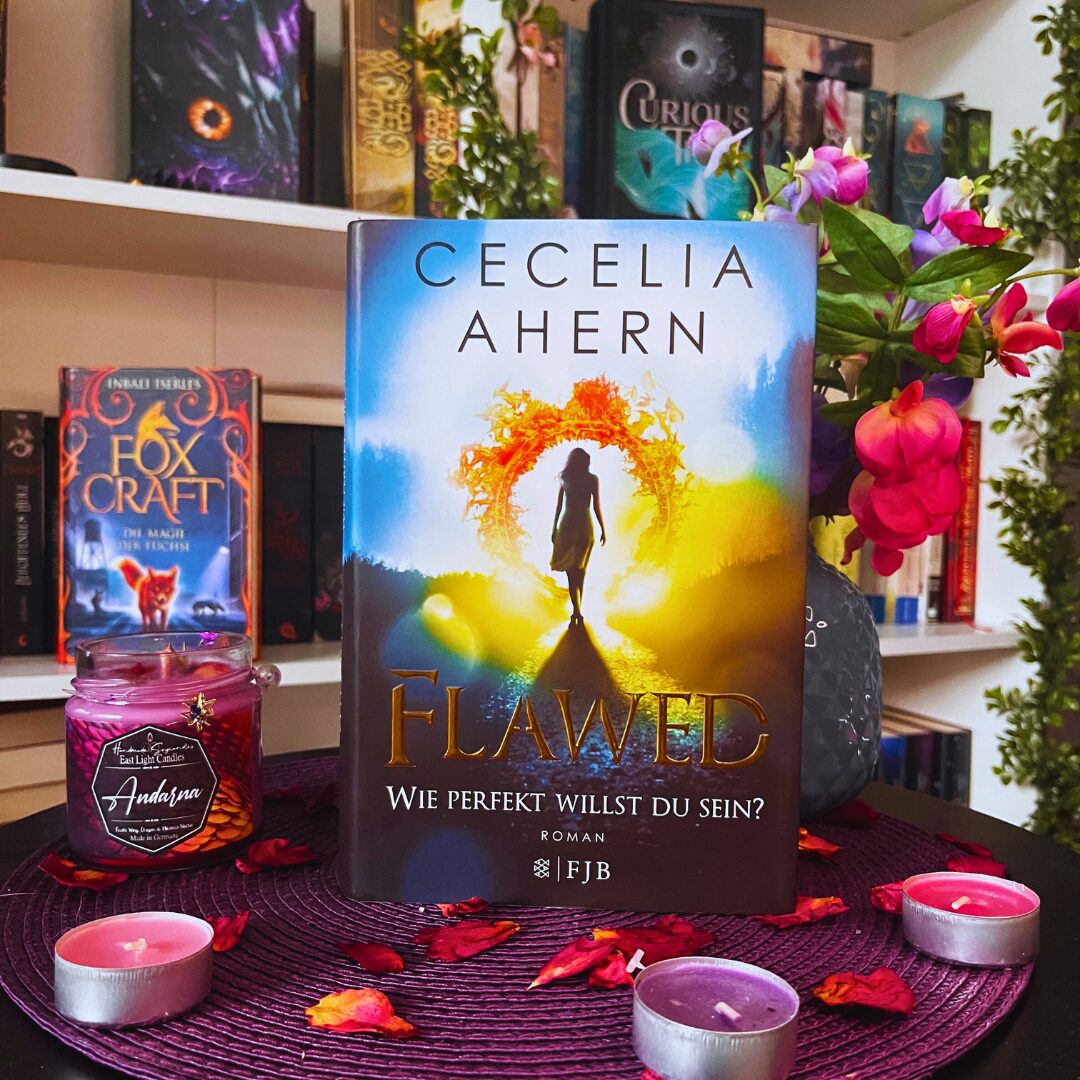 Das Bild zeigt ein Buch mit dem Titel "Flawed" von Cecelia Ahern. Der Untertitel lautet "Wie perfekt willst du sein?". Das Buch steht auf einem runden, lila Untersetzer, umgeben von kleinen Teelichtern und getrockneten Blütenblättern. Im Hintergrund sind weitere Bücher in einem Regal zu sehen, darunter ein Buch mit dem Titel "Fox Craft" und ein weiteres mit dem Titel "Curious". Rechts neben dem Buch steht eine Vase mit bunten Blumen.