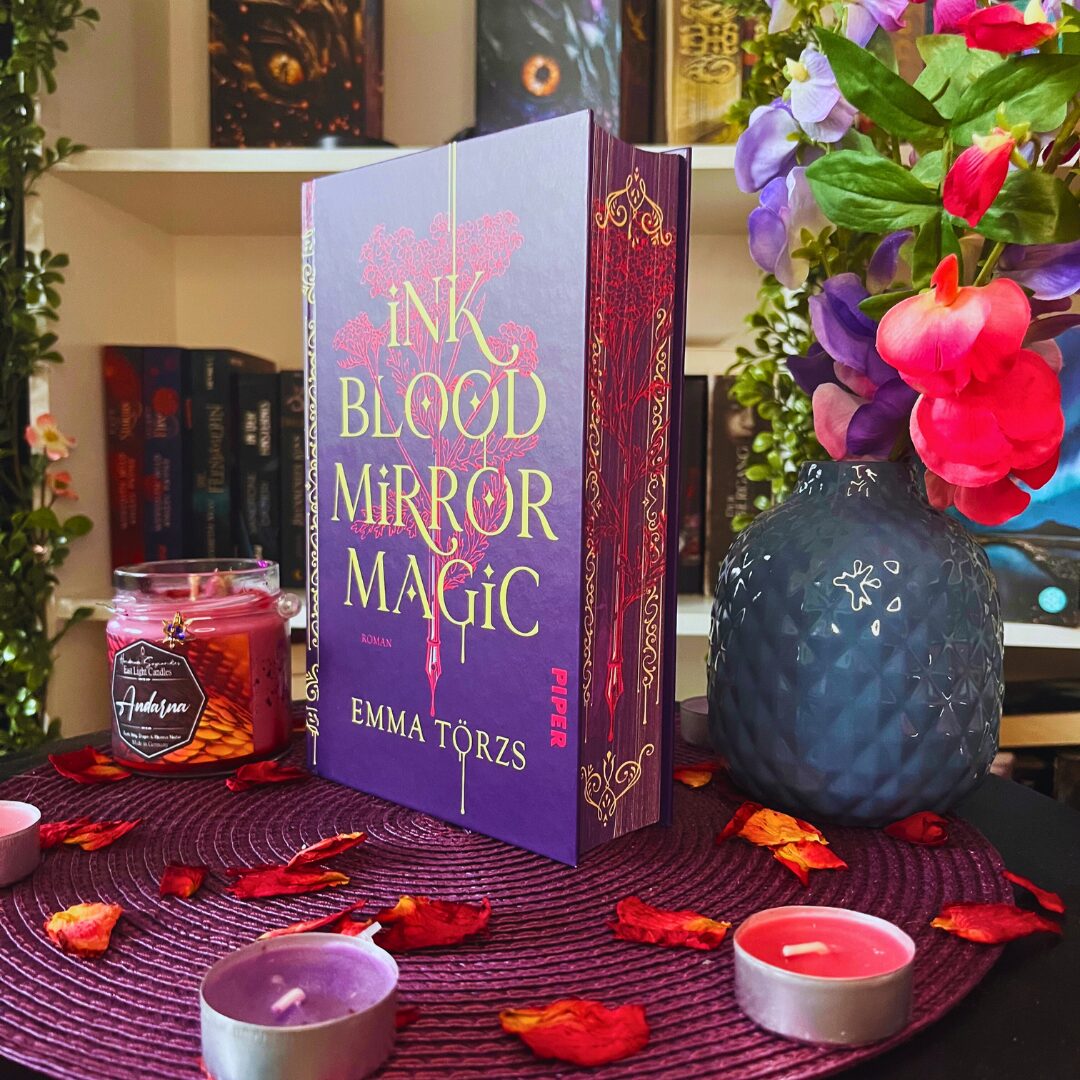 Das Bild zeigt ein Buch mit dem Titel "Ink Blood Mirror Magic" von Emma Törzs. Das Buch hat einen lila Einband mit goldener und roter Schrift. Es steht aufrecht auf einem runden, lila Untersetzer, der auf einem Tisch liegt. Um das Buch herum liegen getrocknete rote Blütenblätter und mehrere kleine Teelichter, von denen einige lila und andere rot sind. Links neben dem Buch steht eine rote Kerze im Glas mit der Aufschrift "Andorra". Rechts vom Buch steht eine dunkelblaue Vase mit einem Muster und darin befinden sich bunte Blumen in Lila, Rosa und Rot. Im Hintergrund sind Bücherregale mit weiteren Büchern zu sehen.