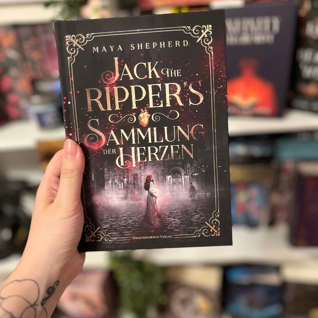 Das Bild zeigt ein Buch, das von einer Hand gehalten wird. Der Titel des Buches lautet "Jack the Ripper's Sammlung der Herzen" und die Autorin ist Maya Shepherd. Das Cover ist überwiegend in dunklen Farben gehalten, mit goldener Schrift für den Titel. Im unteren Teil des Covers ist eine Szene abgebildet, die eine Frau in einem langen Kleid zeigt, die auf einer gepflasterten Straße steht. Im Hintergrund sind alte Gebäude und eine düstere Atmosphäre zu sehen. Das Buch wurde vom Drachenmond Verlag veröffentlicht.