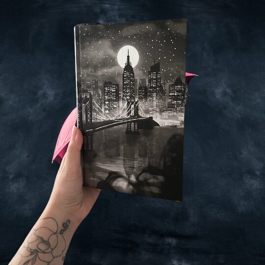 Das Bild zeigt eine Hand, die ein Notizbuch oder Tagebuch hält. Das Cover des Buches hat eine schwarz-weiße Illustration einer Stadt bei Nacht. Im Vordergrund ist eine Brücke zu sehen, die über einen Fluss führt. Im Hintergrund sind hohe Wolkenkratzer und ein großer, runder Vollmond am Himmel zu erkennen. Der Himmel ist mit vielen kleinen Sternen bedeckt. Die Hand, die das Buch hält, hat ein Tattoo eines Blumenmusters auf dem Unterarm. Der Hintergrund des Bildes ist dunkel und unscharf, sodass der Fokus auf dem Buch liegt.