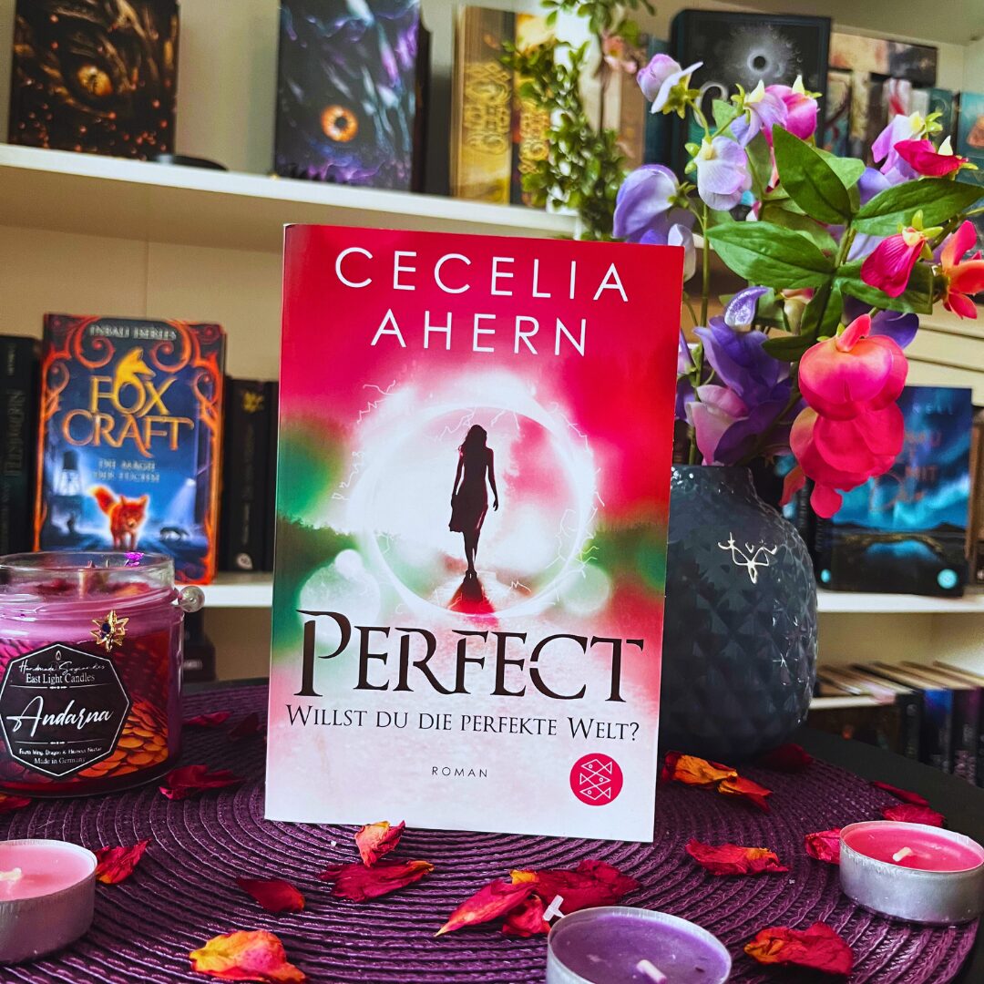 Das Bild zeigt das Buch "Perfect" von Cecelia Ahern. Der Untertitel lautet "Willst du die perfekte Welt?". Das Buch steht auf einem Tisch, der mit einem lila Tuch bedeckt ist. Um das Buch herum liegen getrocknete Blütenblätter und es stehen mehrere Kerzen in verschiedenen Farben, darunter lila und rosa. Im Hintergrund sind Bücherregale mit weiteren Büchern zu sehen. Eines der Bücher im Regal trägt den Titel "Fox Craft". Daneben steht eine Vase mit bunten Blumen.