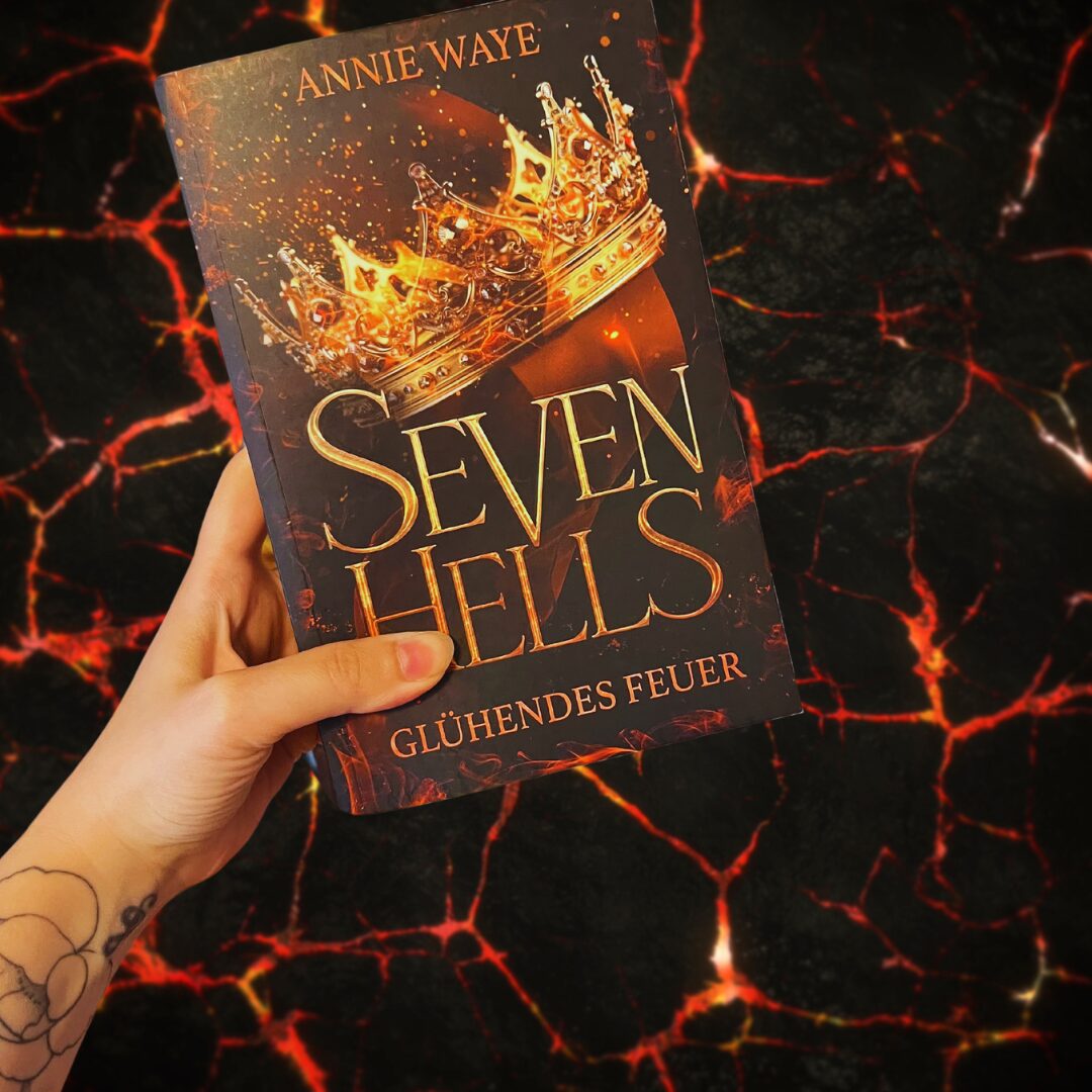 Das Bild zeigt eine Hand, die ein Buch hält. Der Titel des Buches lautet "Seven Hells: Glühendes Feuer" und der Autor ist Annie Waye. Auf dem Buchcover ist eine goldene Krone abgebildet, die von Flammen umgeben ist. Der Hintergrund des Bildes zeigt eine dunkle, feurige Landschaft, die an Lava oder glühende Kohlen erinnert. Die Hand, die das Buch hält, hat ein Tattoo eines Blumenmusters am Handgelenk.