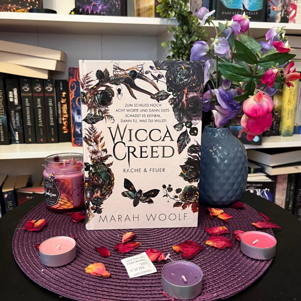 Auf dem Bild ist ein Buch mit dem Titel "Wicca Creed: Rache & Feuer" von Marah Woolf zu sehen. Das Buch steht aufrecht auf einem runden, lila Platzdeckchen. Um das Buch herum liegen getrocknete Blütenblätter und mehrere Teelichter in verschiedenen Farben, darunter lila und rosa. Links neben dem Buch steht eine lila Duftkerze in einem Glas. Rechts neben dem Buch steht eine blaue Vase mit bunten Blumen, hauptsächlich in Lila- und Rosatönen. Im Hintergrund ist ein Bücherregal mit verschiedenen Büchern zu sehen.