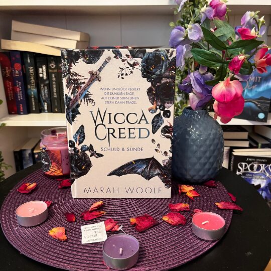 Auf dem Bild ist ein Buch mit dem Titel "Wicca Creed: Rache & Feuer" von Marah Woolf zu sehen. Das Buch steht aufrecht auf einem runden, lila Platzdeckchen. Um das Buch herum liegen getrocknete Blütenblätter und mehrere Teelichter in verschiedenen Farben, darunter lila und rosa. Links neben dem Buch steht eine lila Duftkerze in einem Glas. Rechts neben dem Buch steht eine blaue Vase mit bunten Blumen, hauptsächlich in Lila- und Rosatönen. Im Hintergrund ist ein Bücherregal mit verschiedenen Büchern zu sehen.