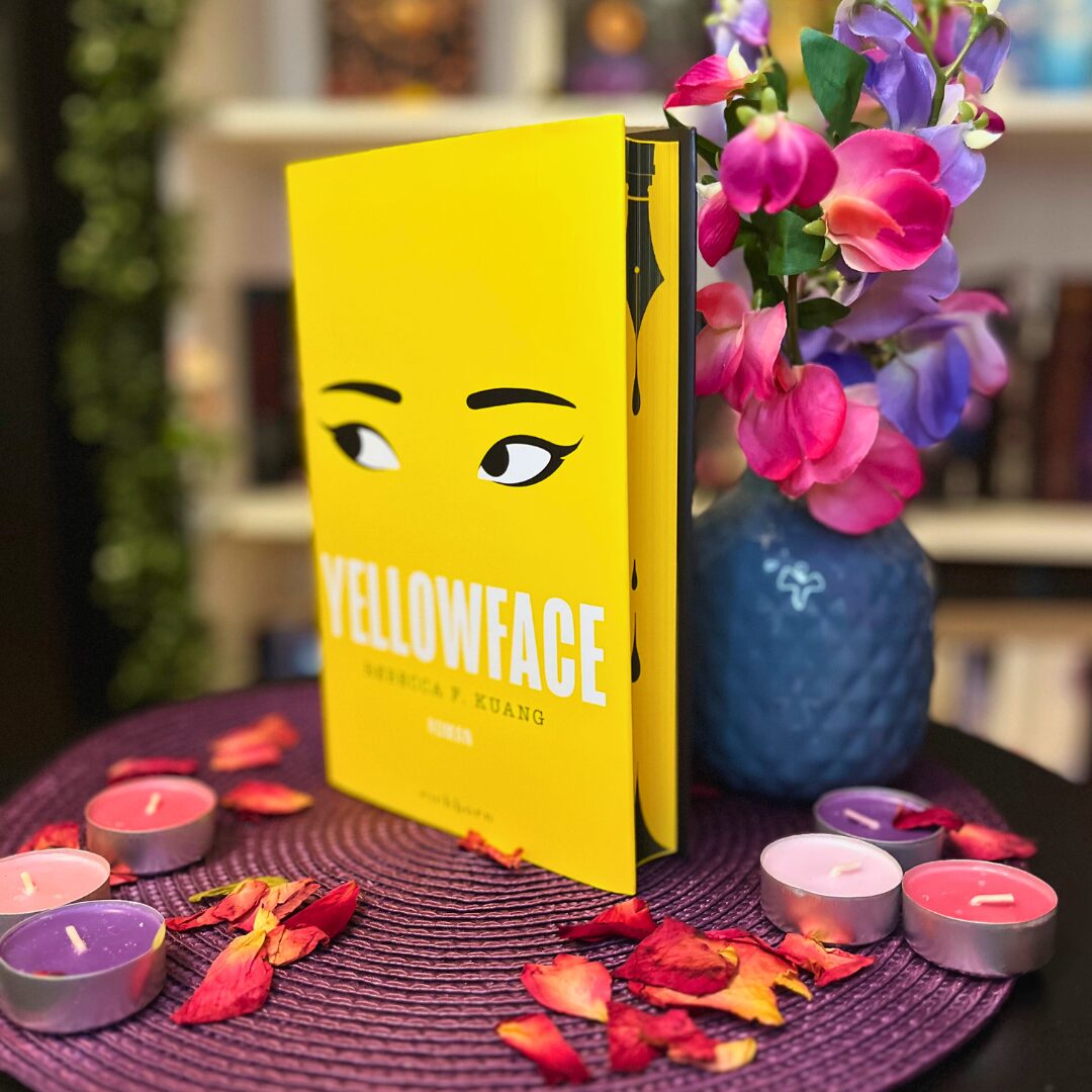 Das Bild zeigt ein Buch mit dem Titel "Yellowface" von Rebecca F. Kuang. Das Buch hat einen leuchtend gelben Einband mit einer Illustration von Augen, die nach rechts schauen. Es steht auf einem runden, lila Untersetzer, der auf einem Tisch liegt. Um das Buch herum sind mehrere kleine Teelichter in verschiedenen Farben (rosa, lila und rot) sowie verstreute Rosenblätter. Rechts neben dem Buch steht eine blaue Vase mit bunten Blumen in Rosa, Lila und Violett. Im Hintergrund sind unscharf Bücherregale zu erkennen.