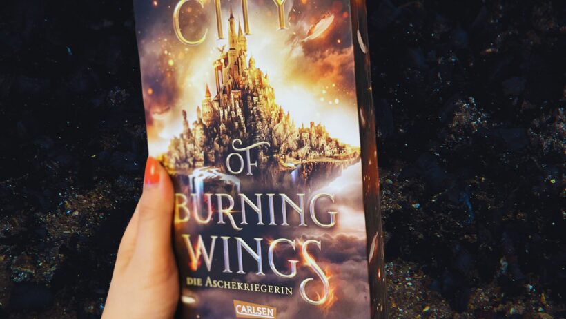 Das Bild zeigt ein Buch, das von einer Hand gehalten wird. Der Titel des Buches lautet "City of Burning Wings: Die Aschekriegerin" und es ist von der Autorin Lily S. Morgan. Auf dem Cover ist eine fantasievolle Stadt abgebildet, die auf einem Felsen zu schweben scheint, umgeben von einem dramatischen Himmel mit leuchtenden Wolken und funkelnden Lichtern. Die Hand, die das Buch hält, hat ein Tattoo mit einem Blumenmotiv auf dem Handgelenk. Der Hintergrund des Bildes ist dunkel und unscharf, was das Buch in den Vordergrund rückt.