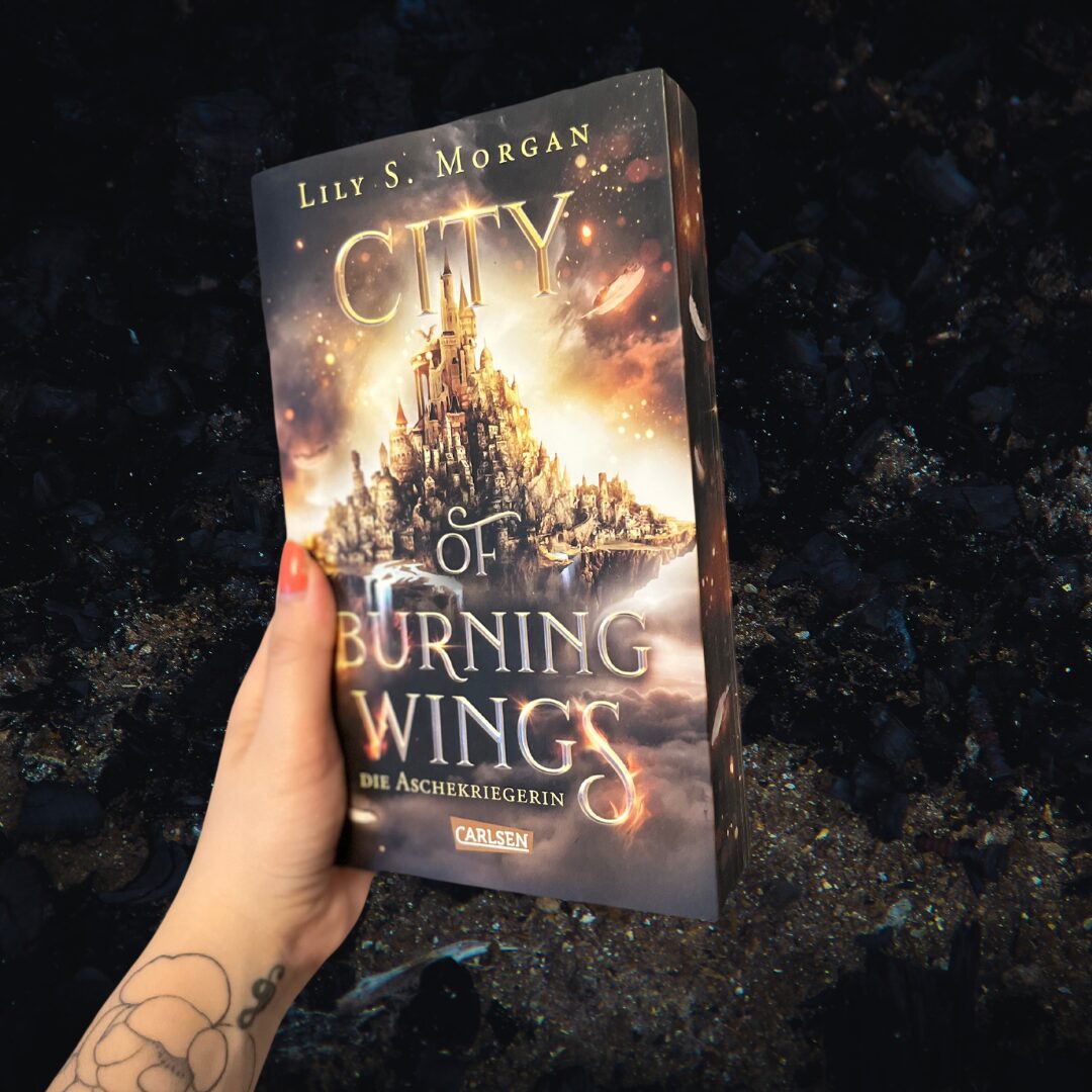 Das Bild zeigt ein Buch, das von einer Hand gehalten wird. Der Titel des Buches lautet "City of Burning Wings: Die Aschekriegerin" und es ist von der Autorin Lily S. Morgan. Auf dem Cover ist eine fantasievolle Stadt abgebildet, die auf einem Felsen zu schweben scheint, umgeben von einem dramatischen Himmel mit leuchtenden Wolken und funkelnden Lichtern. Die Hand, die das Buch hält, hat ein Tattoo mit einem Blumenmotiv auf dem Handgelenk. Der Hintergrund des Bildes ist dunkel und unscharf, was das Buch in den Vordergrund rückt.