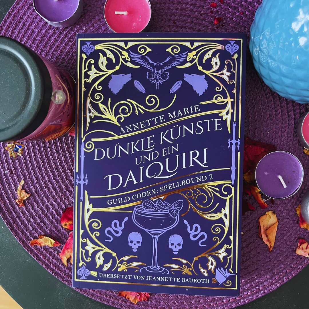 Das Bild zeigt ein Buch mit dem Titel "Dunkle Künste und ein Daiquiri" von Annette Marie. Es ist der zweite Band der Reihe "Guild Codex: Spellbound". Das Cover ist in dunklem Lila gehalten und mit goldenen Verzierungen geschmückt. Es zeigt ein Cocktailglas mit einem Daiquiri und zwei Totenköpfen darunter. Um das Buch herum liegen verschiedene Kerzen in Lila und Rot sowie getrocknete Blütenblätter auf einem lila Tischset.