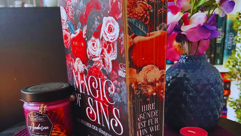 Das Bild zeigt ein Buch mit dem Titel "Magic of Sins" von Caroline Oiseau. Das Cover ist reich verziert mit roten und weißen Rosen sowie anderen Blumen. Neben dem Buch steht ein Glas mit einer lila Kerze, auf dem "Andorra" steht. Im Hintergrund ist eine dunkelblaue Vase mit lila und rosa Blumen zu sehen. Auf dem Tisch liegen einige getrocknete Blütenblätter und es gibt auch ein paar kleine Teelichter. Alles steht auf einer lila Unterlage.