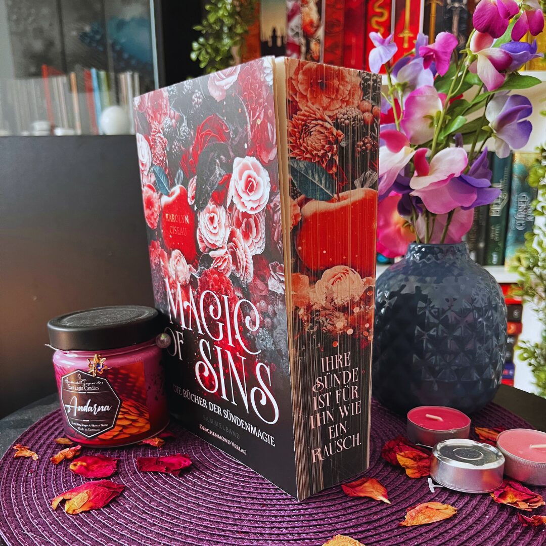 Das Bild zeigt ein Buch mit dem Titel "Magic of Sins" von Caroline Oiseau. Das Cover ist reich verziert mit roten und weißen Rosen sowie anderen Blumen. Neben dem Buch steht ein Glas mit einer lila Kerze, auf dem "Andorra" steht. Im Hintergrund ist eine dunkelblaue Vase mit lila und rosa Blumen zu sehen. Auf dem Tisch liegen einige getrocknete Blütenblätter und es gibt auch ein paar kleine Teelichter. Alles steht auf einer lila Unterlage. 