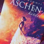 Das Bild zeigt ein Buch mit dem Titel "Aschenkindel" von Halo Summer. Auf dem Cover ist eine Frau in einem eleganten, weißen Kleid abgebildet, die in einem warmen, leuchtenden Licht steht. Der Hintergrund hat eine feurige, märchenhafte Atmosphäre. Das Buch liegt auf einem lila, geflochtenen Untersetzer, umgeben von mehreren kleinen, bunten Kerzen und getrockneten Blättern. Unten auf dem Buchcover steht "Das wahre Märchen" und "Drachenmond Verlag".