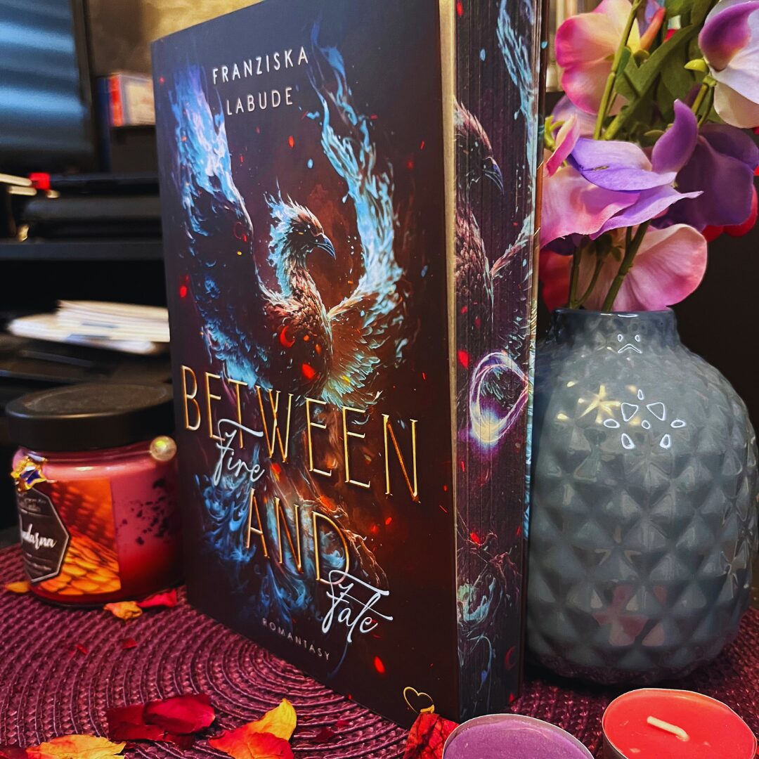 Das Bild zeigt ein Buch mit dem Titel "Between Fire and Fate" von Franziska Labude. Auf dem Buchcover ist ein majestätischer Vogel abgebildet, der von Flammen und blauen Lichtern umgeben ist. Das Buch steht auf einem Tisch, der mit einem lila Platzdeckchen bedeckt ist. Daneben befinden sich eine rote Kerze in einem Glas mit einem Etikett und ein paar bunte Blütenblätter. Rechts vom Buch steht eine Vase mit lila und rosa Blumen. Im Hintergrund sind einige unscharfe Gegenstände zu sehen, darunter ein Stapel Papier.