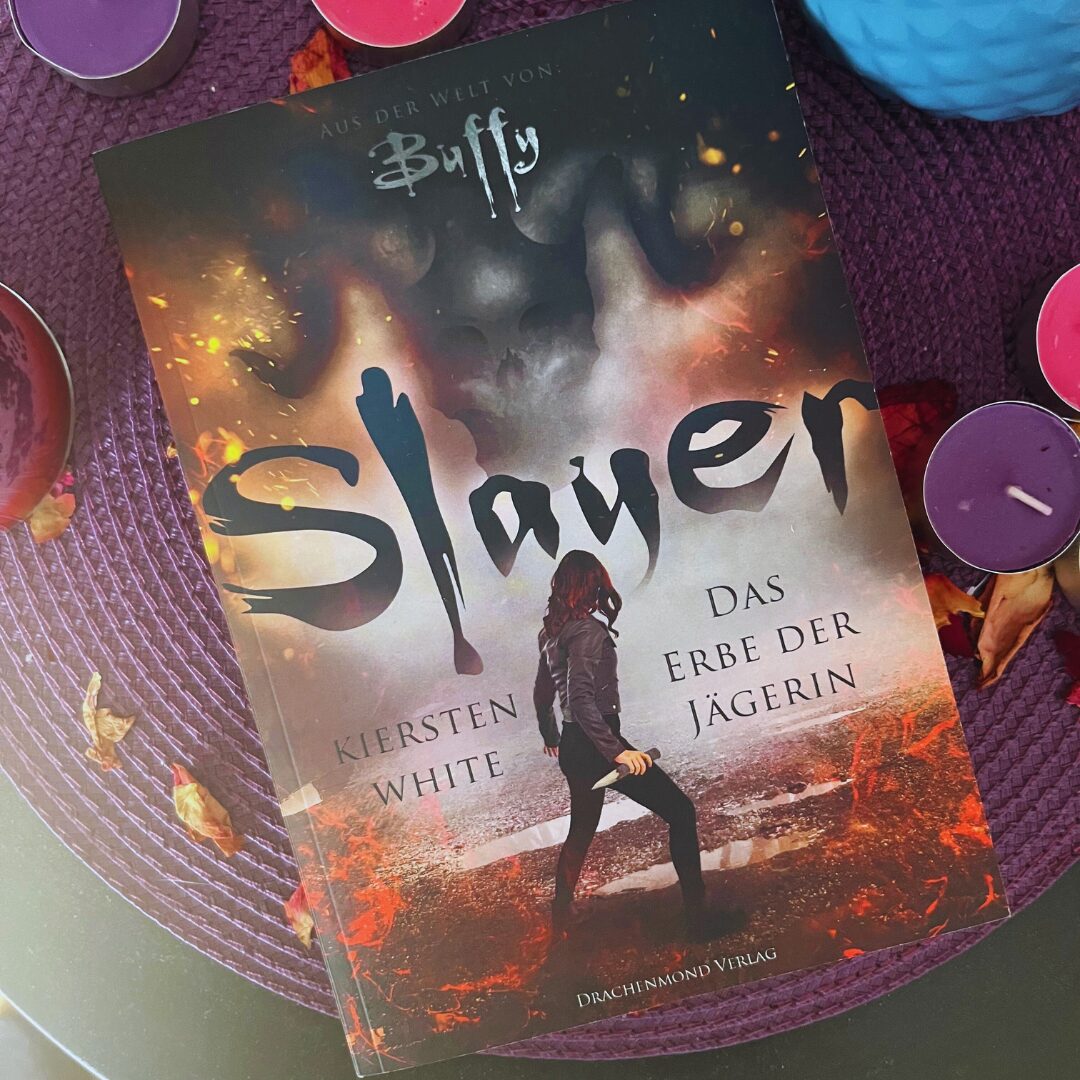 Das Bild zeigt das Cover eines Buches mit dem Titel "Slayer: Das Erbe der Jägerin" von Kiersten White. Es ist aus der Welt von "Buffy". Auf dem Cover ist eine Person mit langen Haaren und dunkler Kleidung abgebildet, die in einer feurigen und rauchigen Umgebung steht. Der Hintergrund ist dramatisch und düster gestaltet. Das Buch liegt auf einem lila Untergrund, umgeben von mehreren kleinen, bunten Kerzen und einigen Herbstblättern.