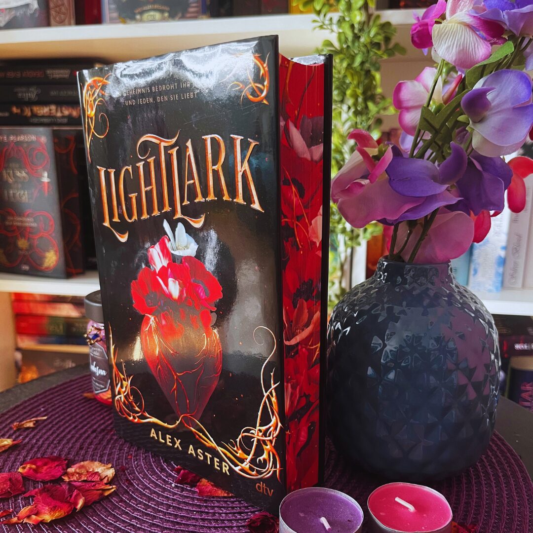 Das Bild zeigt ein Buch mit dem Titel "Lightlark" von Alex Aster. Das Cover ist überwiegend schwarz mit einem auffälligen roten Design, das eine Art Herz oder Blume darstellt, umgeben von dekorativen, goldenen Ranken. Der Buchrücken ist ebenfalls rot mit floralen Mustern. Das Buch steht auf einem Tisch, der mit einem lila Platzdeckchen bedeckt ist. Daneben liegt eine Vase mit lila und rosa Blumen. Vor dem Buch stehen zwei kleine Kerzen, eine in lila und eine in pink. Im Hintergrund sind Bücherregale mit weiteren Büchern zu sehen.