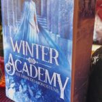 Das Bild zeigt ein Buch mit dem Titel "Winter Academy: Die Erbin des Mondsteins" von Karolyn Ciseau. Das Cover ist in kühlen Blautönen gehalten und zeigt eine Frau in einem blauen Kleid, die vor einem Torbogen steht. Im Hintergrund sind Eiskristalle und winterliche Motive zu sehen. Das Buch steht auf einem Tisch, der mit roten Blütenblättern dekoriert ist. Daneben steht eine Vase mit rosa und lila Blumen. Im Hintergrund ist ein Teil einer Pflanze zu erkennen.