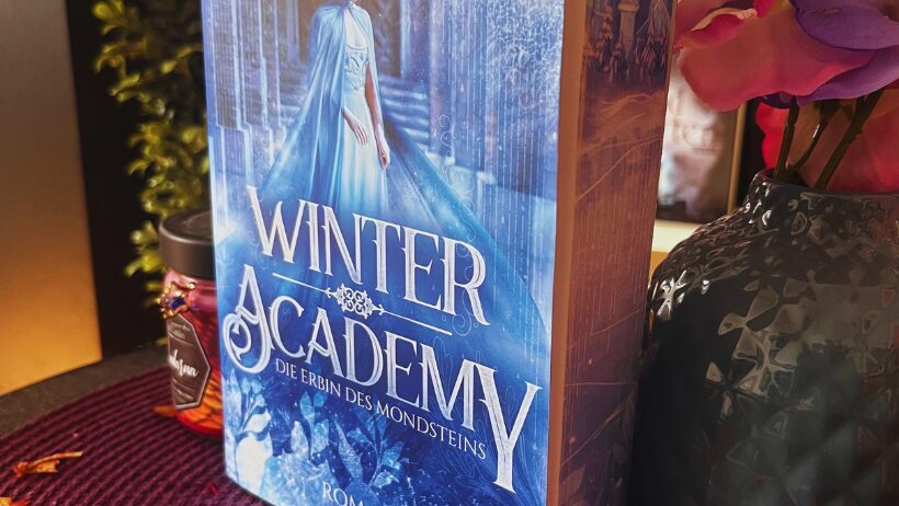 Das Bild zeigt ein Buch mit dem Titel "Winter Academy: Die Erbin des Mondsteins" von Karolyn Ciseau. Das Cover ist in kühlen Blautönen gehalten und zeigt eine Frau in einem blauen Kleid, die vor einem Torbogen steht. Im Hintergrund sind Eiskristalle und winterliche Motive zu sehen. Das Buch steht auf einem Tisch, der mit roten Blütenblättern dekoriert ist. Daneben steht eine Vase mit rosa und lila Blumen. Im Hintergrund ist ein Teil einer Pflanze zu erkennen.