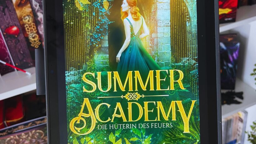 Das Bild zeigt ein Tablet, auf dem das Cover eines Buches angezeigt wird. Der Titel des Buches ist "Summer Academy: Die Hüterin des Feuers" von Karolyn Ciseau. Auf dem Cover ist eine Frau mit langen roten Haaren in einem eleganten, grünen Kleid abgebildet. Sie steht vor einem steinernen Torbogen, der von Pflanzen und leuchtenden Lichtern umgeben ist. Im Hintergrund sind Bäume und ein mystischer Wald zu sehen. Das Cover hat eine märchenhafte und magische Atmosphäre. Im Hintergrund des Bildes sind Bücherregale mit weiteren Büchern zu erkennen.