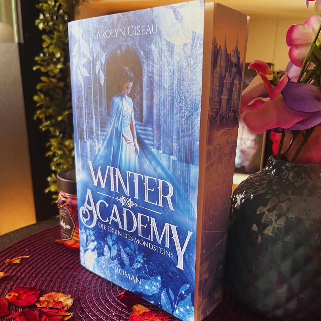 Das Bild zeigt ein Buch mit dem Titel "Winter Academy: Die Erbin des Mondsteins" von Karolyn Ciseau. Das Cover ist in kühlen Blautönen gehalten und zeigt eine Frau in einem blauen Kleid, die vor einem Torbogen steht. Im Hintergrund sind Eiskristalle und winterliche Motive zu sehen. Das Buch steht auf einem Tisch, der mit roten Blütenblättern dekoriert ist. Daneben steht eine Vase mit rosa und lila Blumen. Im Hintergrund ist ein Teil einer Pflanze zu erkennen.